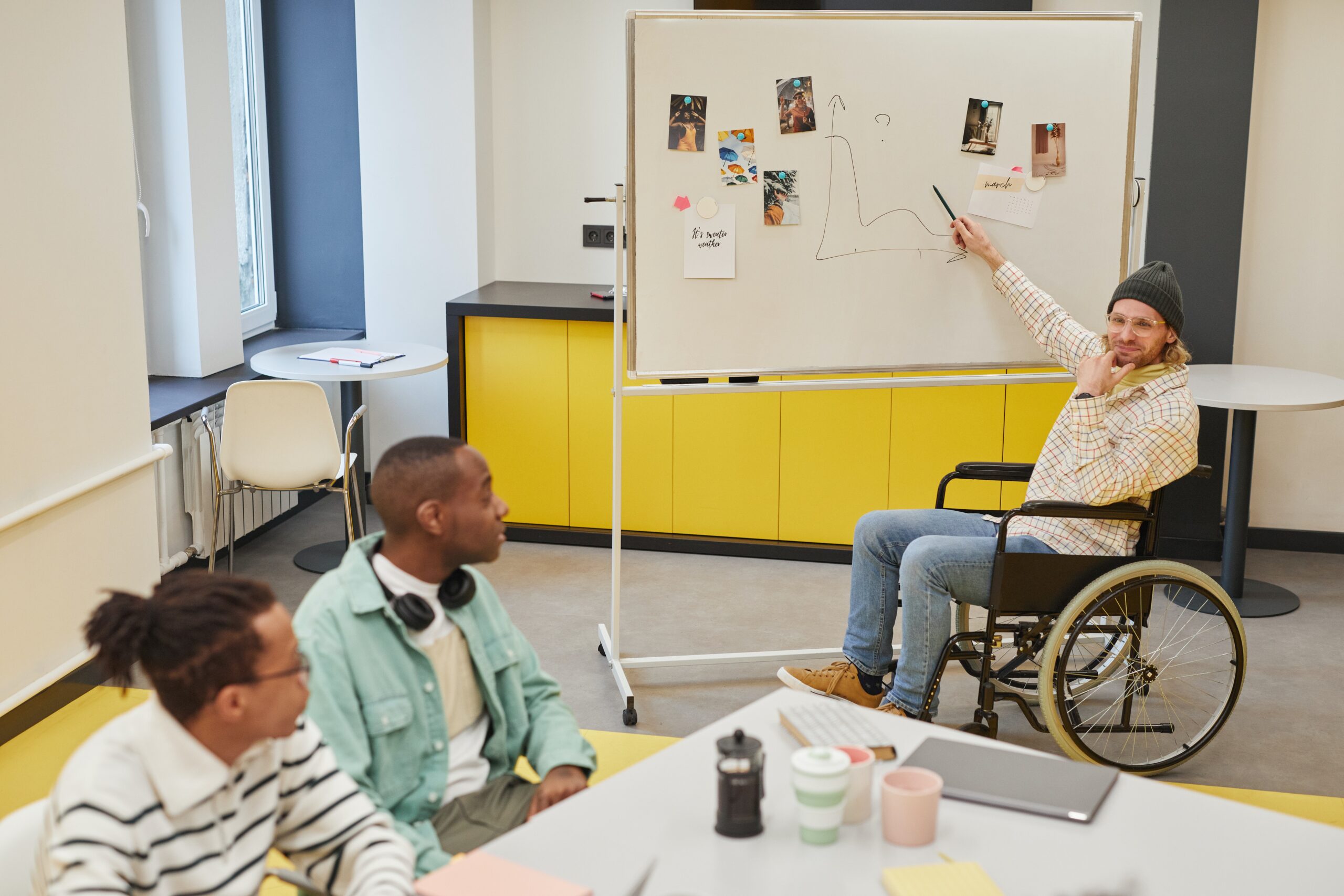 A person in a wheelchair points at a whiteboard while two people look on from their desk.