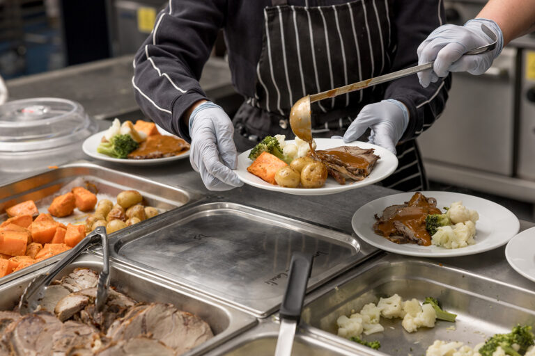 A person in a striped apron and gloves is serving food onto a plate from a buffet, adding gravy to a dish with roasted vegetables and meat.